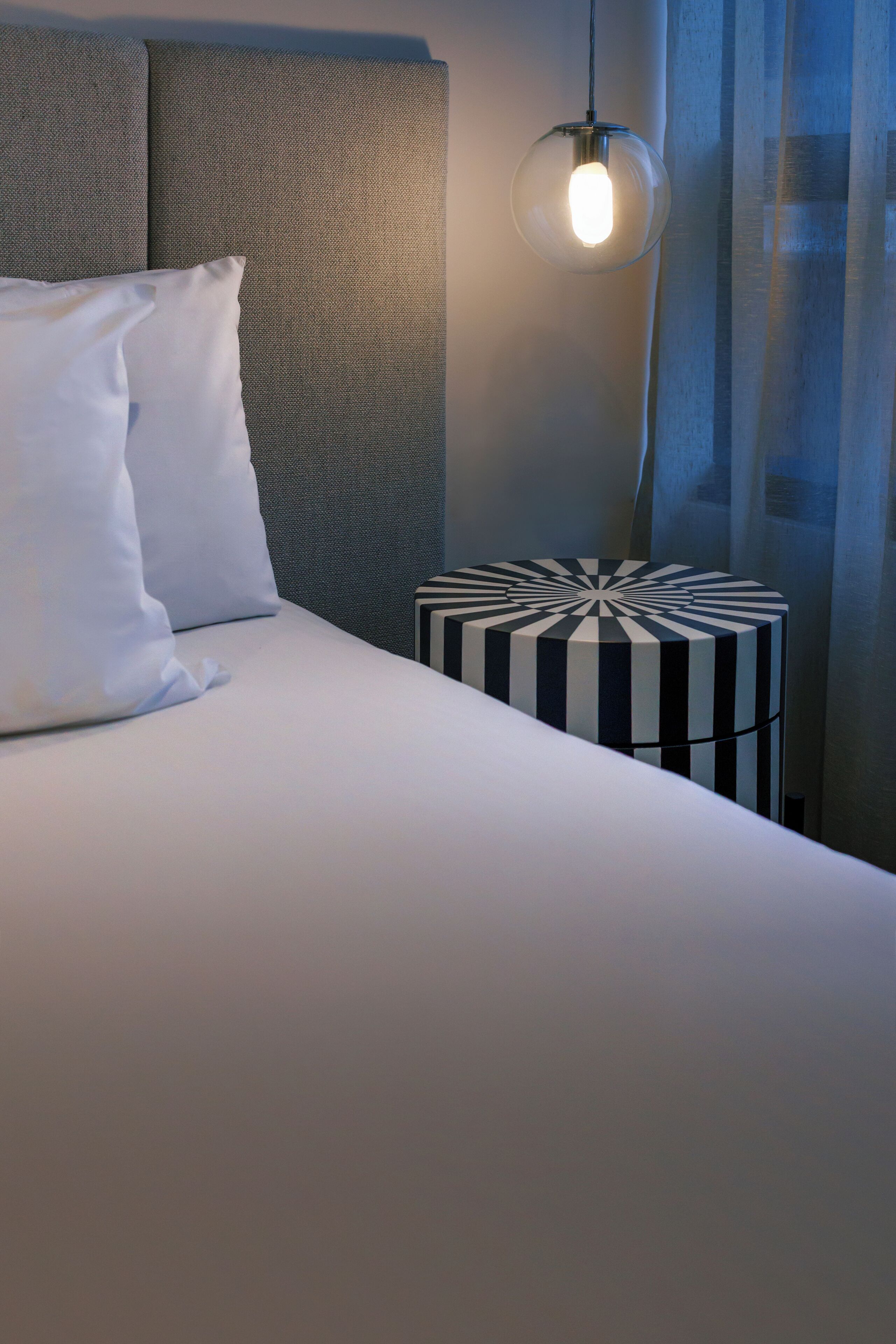 A neatly made bed with white bedding and pillows, a round side table with a striped pattern, and a hanging light fixture.