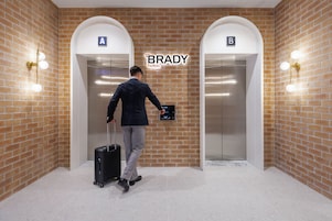 A man in a suit with a suitcase in an elevator with 'BRADY hotels' signage, brick walls, and two elevator doors labeled 'A' and 'B'.