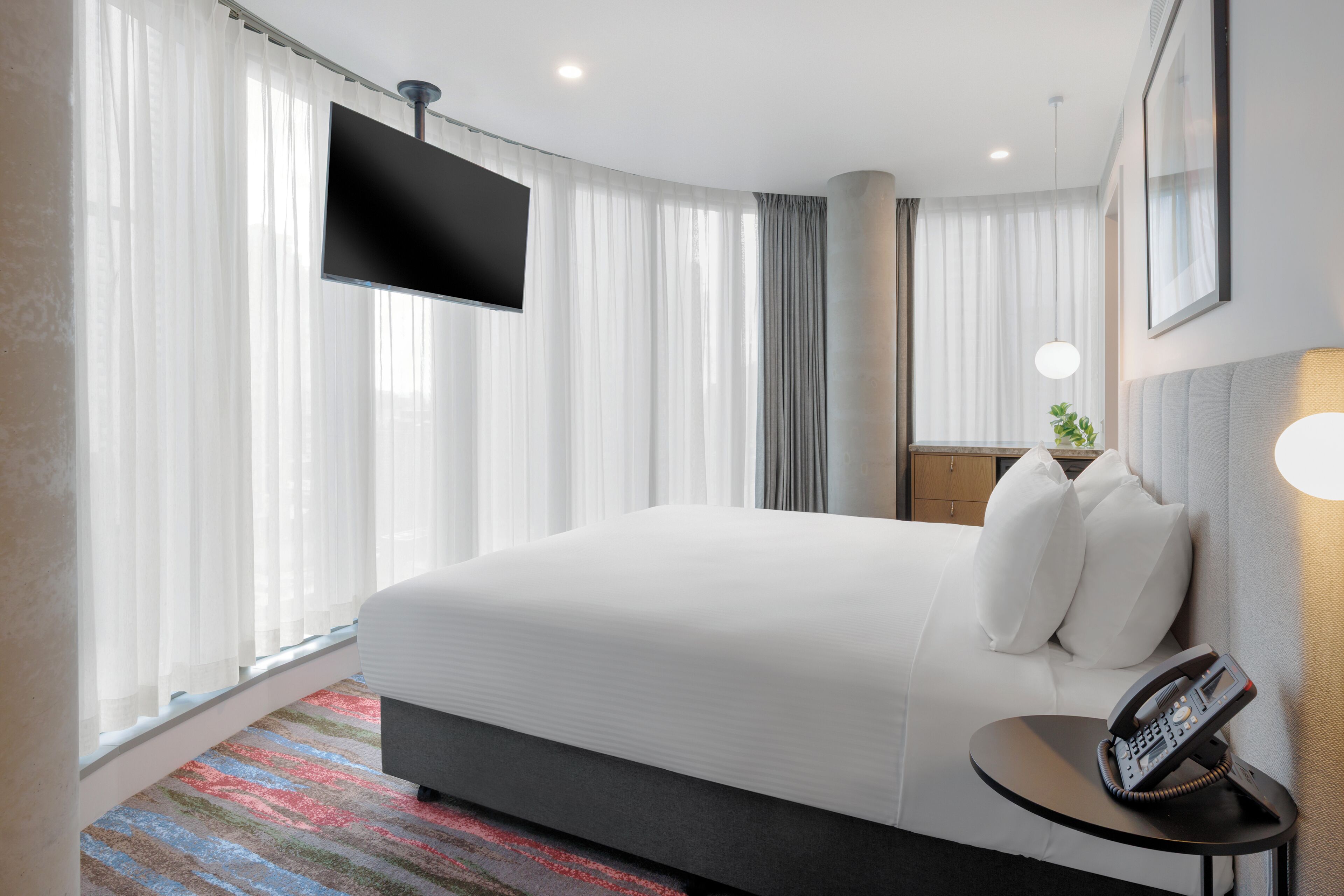 A hotel room with a bed, a TV mounted on the wall, a bedside table with a phone, and a window with curtains.