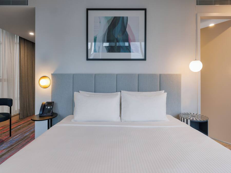 A hotel room with a bed, a bedside table with a phone, a wall-mounted light, and a framed abstract artwork.
