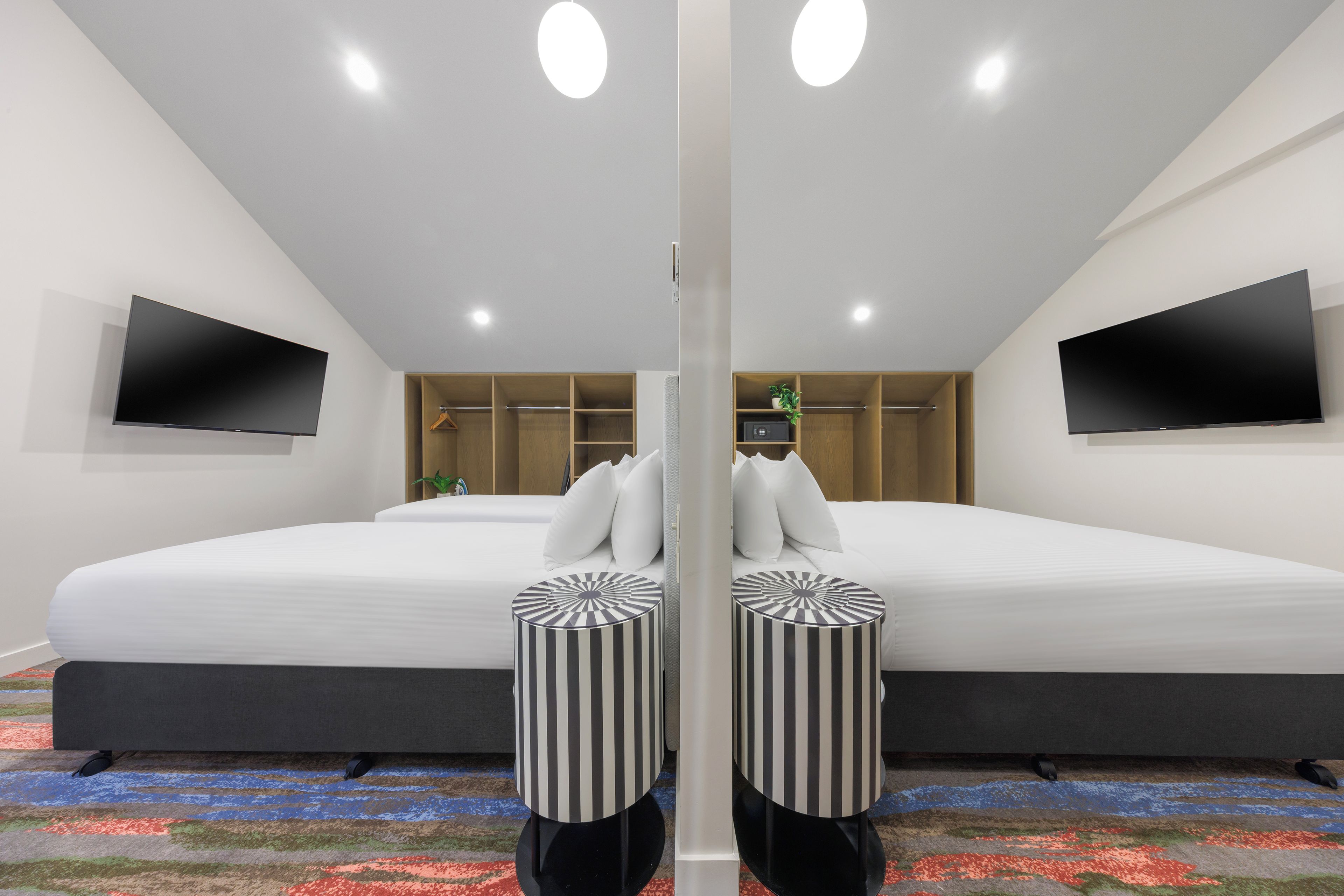 A modern hotel room with a large bed, two side-by-side televisions, a wardrobe, and a small plant.