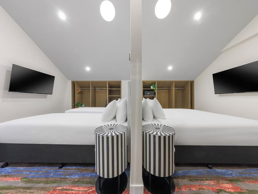 A modern hotel room with a large bed, two side-by-side televisions, a wardrobe, and a small plant.
