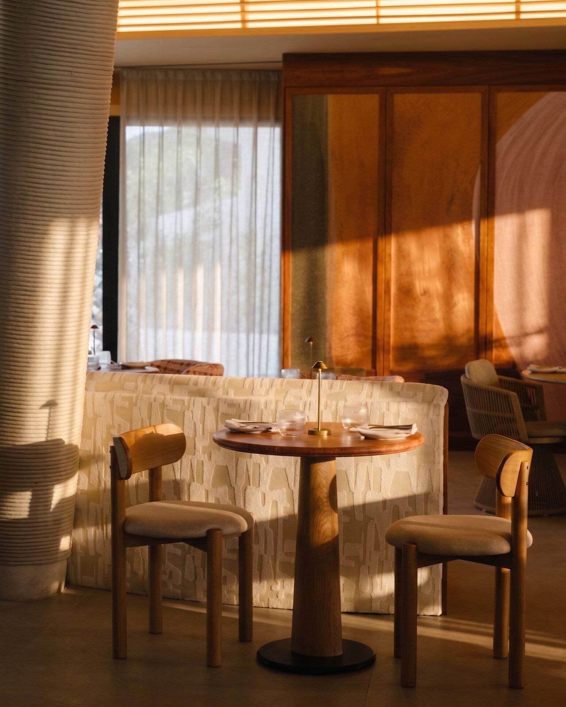 A modern living room with a round wooden table, two wooden chairs, a sofa, and a large window with blinds.