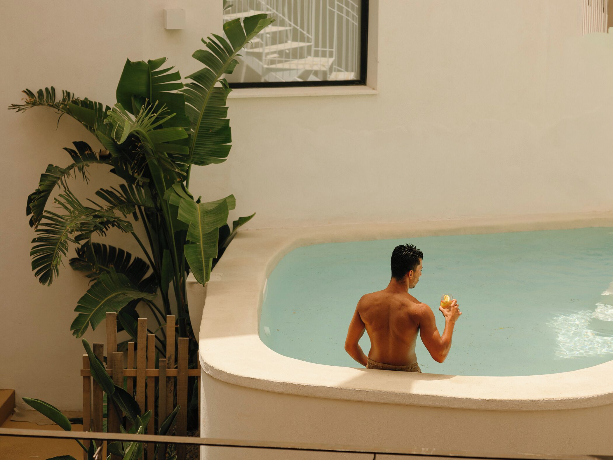 Un hombre en una bañera, con una bebida, rodeado de plantas verdes grandes.