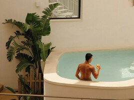 Un hombre en una bañera, con una bebida, rodeado de plantas verdes grandes.