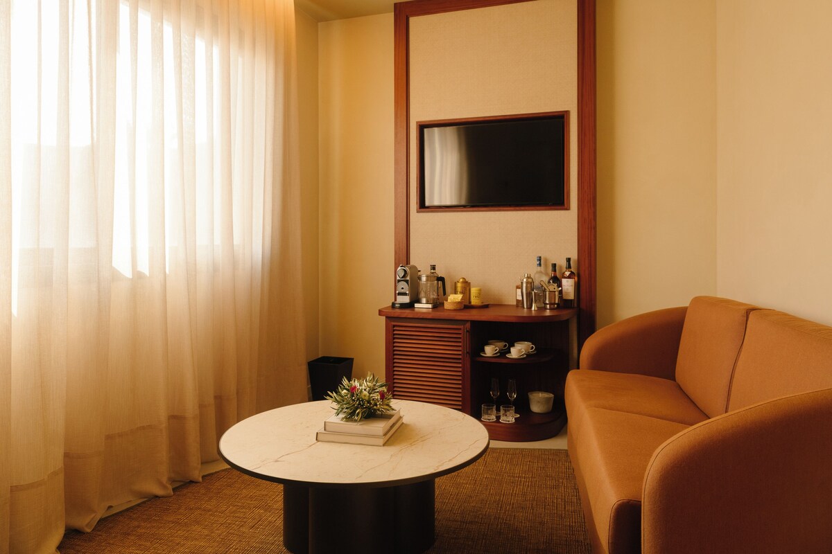 A hotel room with a round table, a sofa, a TV mounted on the wall, and a window with curtains.