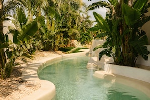 Una piscina de borde curvo, rodeada de plantas tropicales y un muro de piedra.