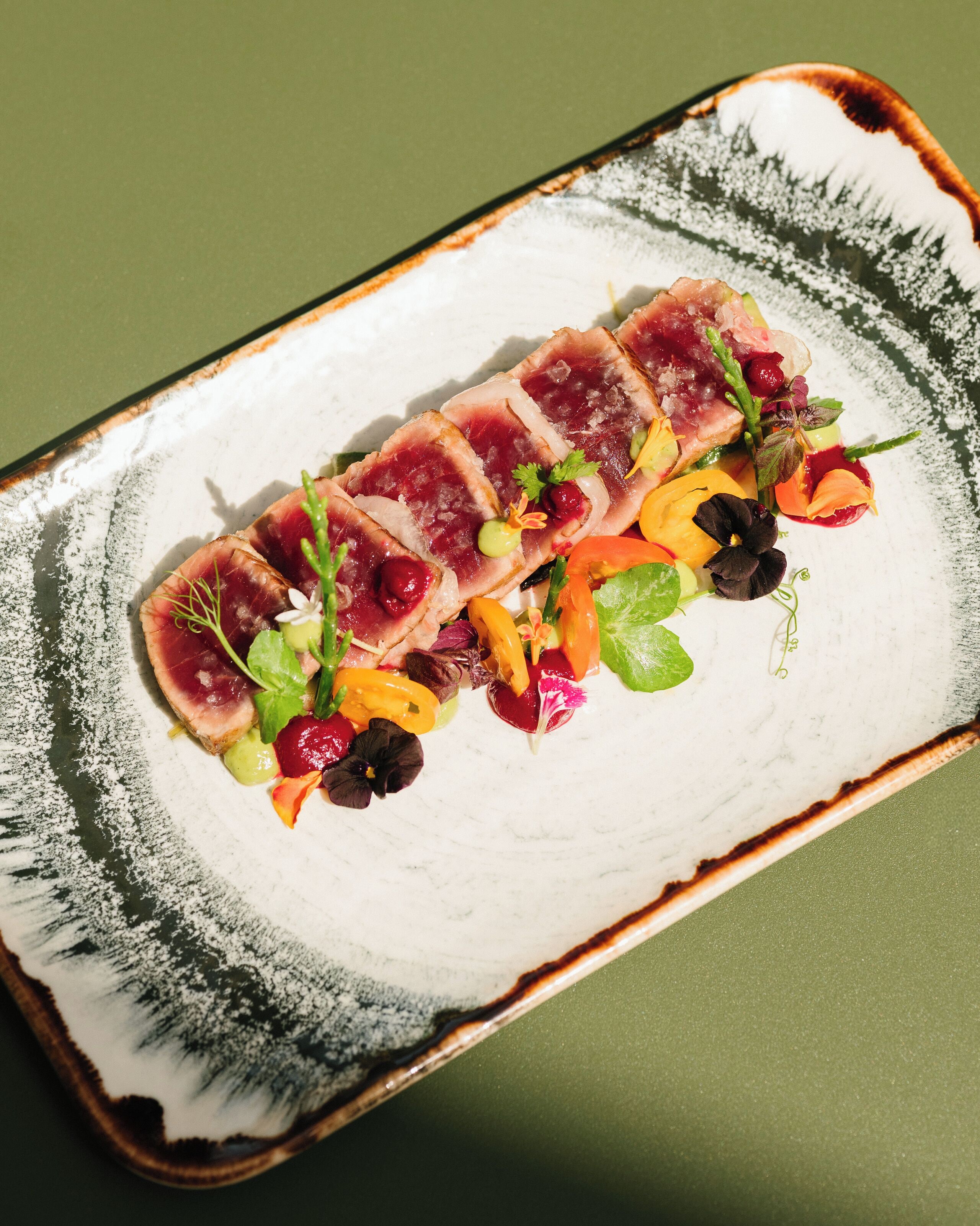 Un plato rectangular con un borde blanco y marrón, que contiene carne en lonchas finas, diversos vegetales y adornos.