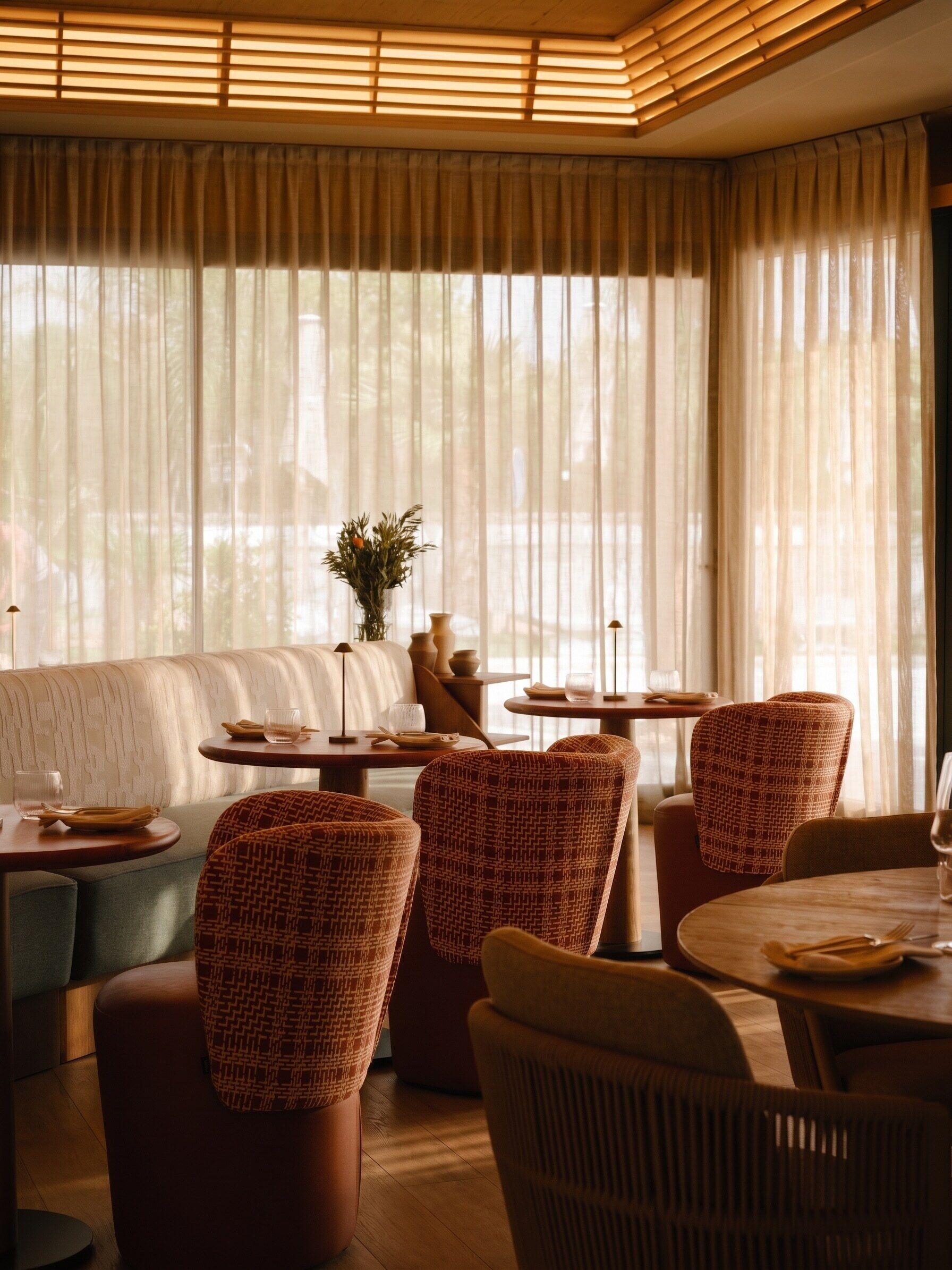 Un restaurante con techo alto, amplios ventanales con cortinas translúcidas y un área de estar con sillas estampadas y un sofá.