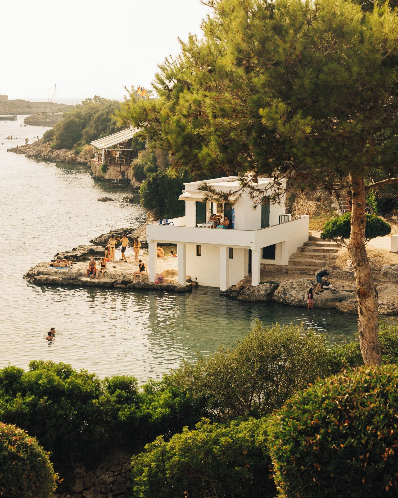 A white building with a balcony overlooking a body of water, surrounded by greenery.