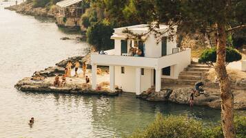 A white building with a balcony overlooking a body of water, surrounded by greenery.