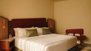 Quarto de hotel com uma cama grande, cabeceira de madeira, duas mesas de cabeceira e um banquinho vermelho.
