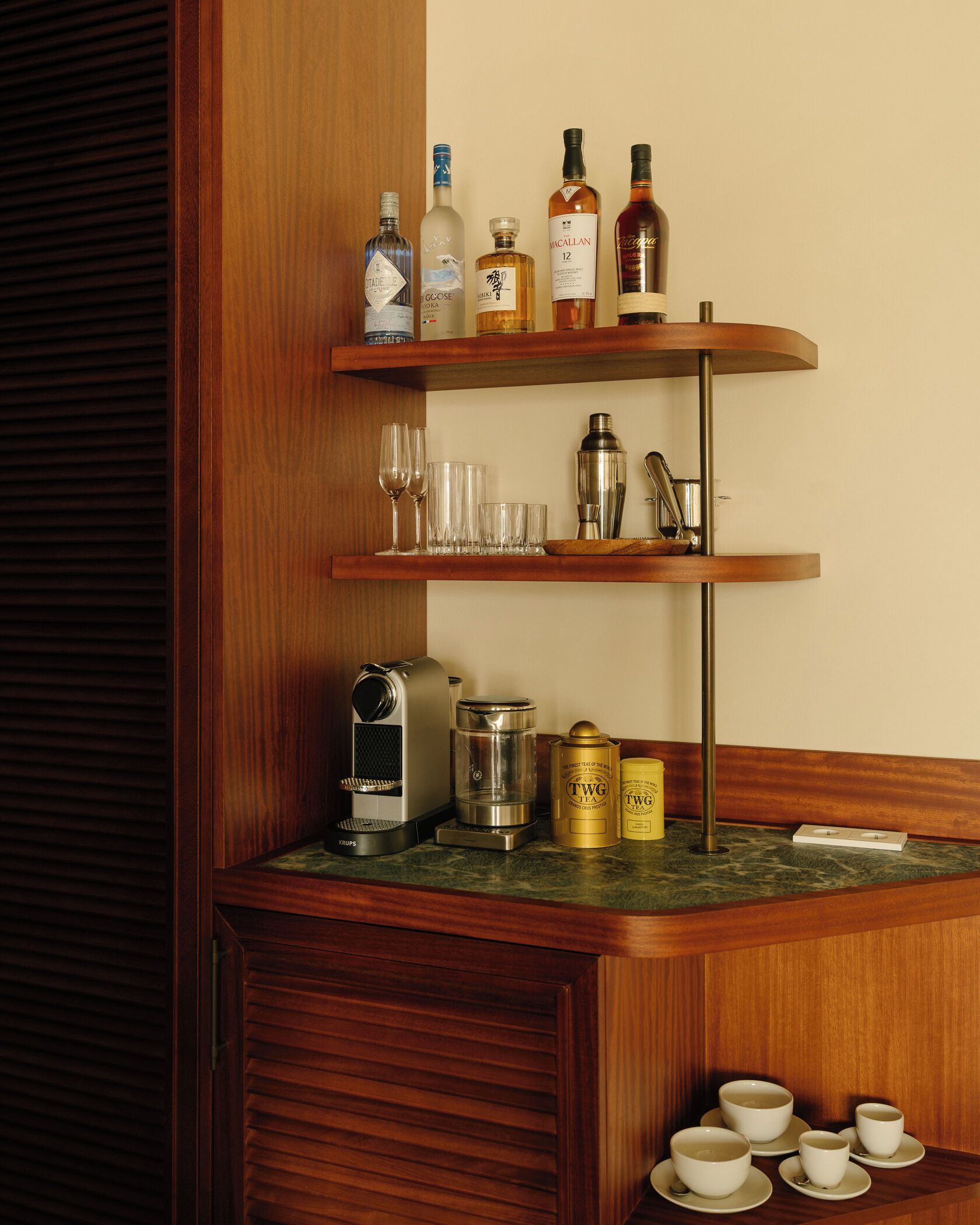 Una encimera de cocina con una cafetera, latas de té y una estantería con botellas y vasos.
