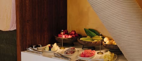 Een buffet met diverse soorten fruit en groenten, waaronder appels, bananen en watermeloen, plus sauzen en bestek.