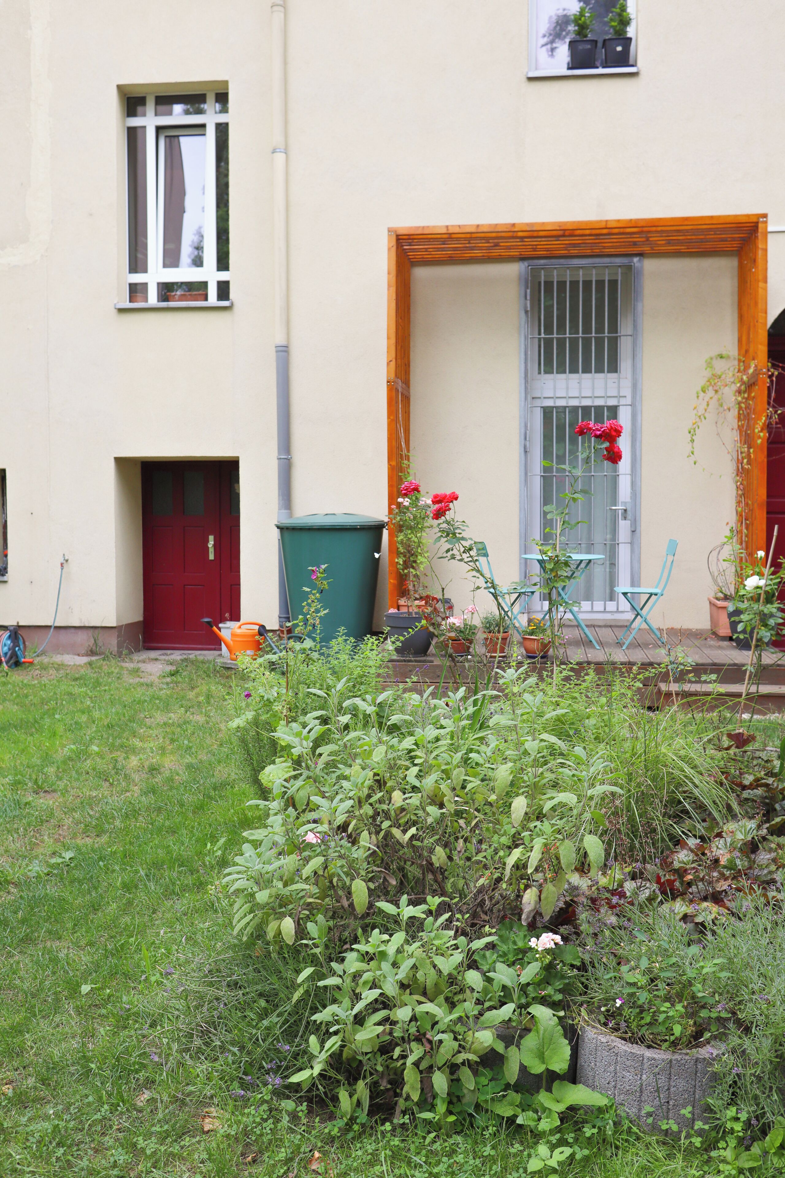 A residential building with a red door, a window with curtains, a wooden-framed entrance, and a small garden area with potted plants and a table.