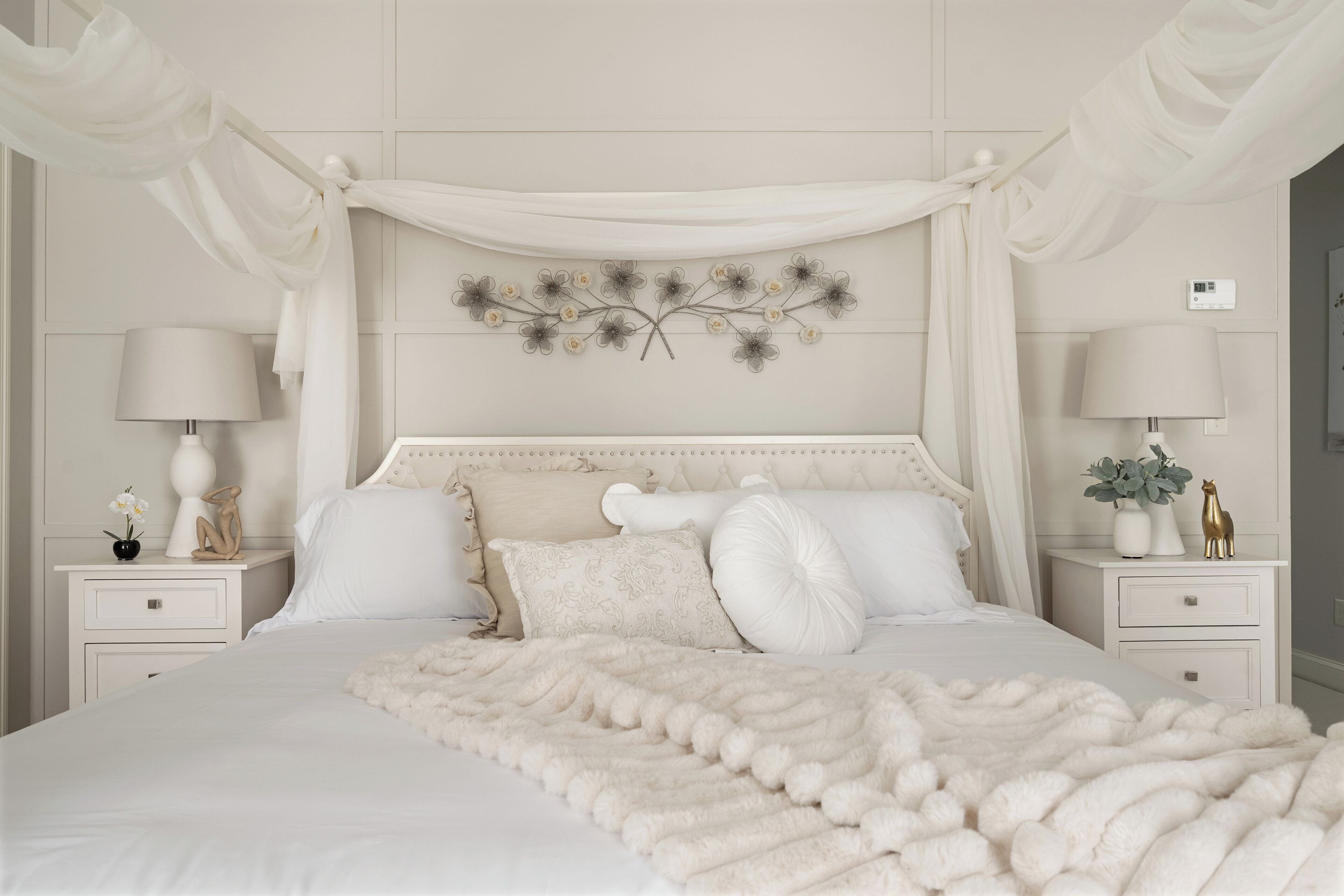 A neatly made bed with a fluffy white blanket, a decorative wall piece, and a white canopy.