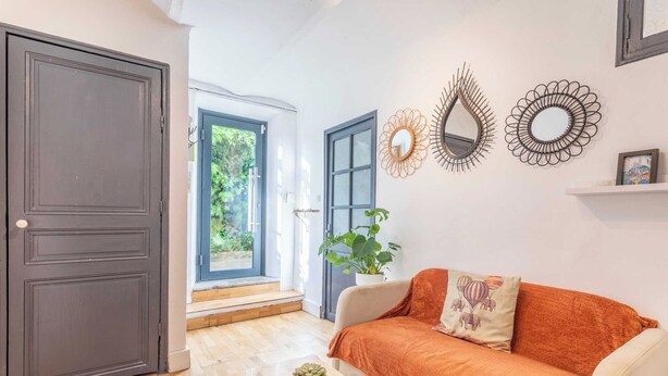 Ein Zimmer mit Holzboden, einem Sofa mit orangefarbener Decke, einer Topfpflanze und dekorativen Wandspiegeln.