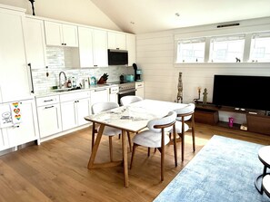 A modern kitchen with white cabinets, a dining table with four chairs, a TV, and a window with blinds. - Modern West End Retreat with 2 King Beds (Provincetown)