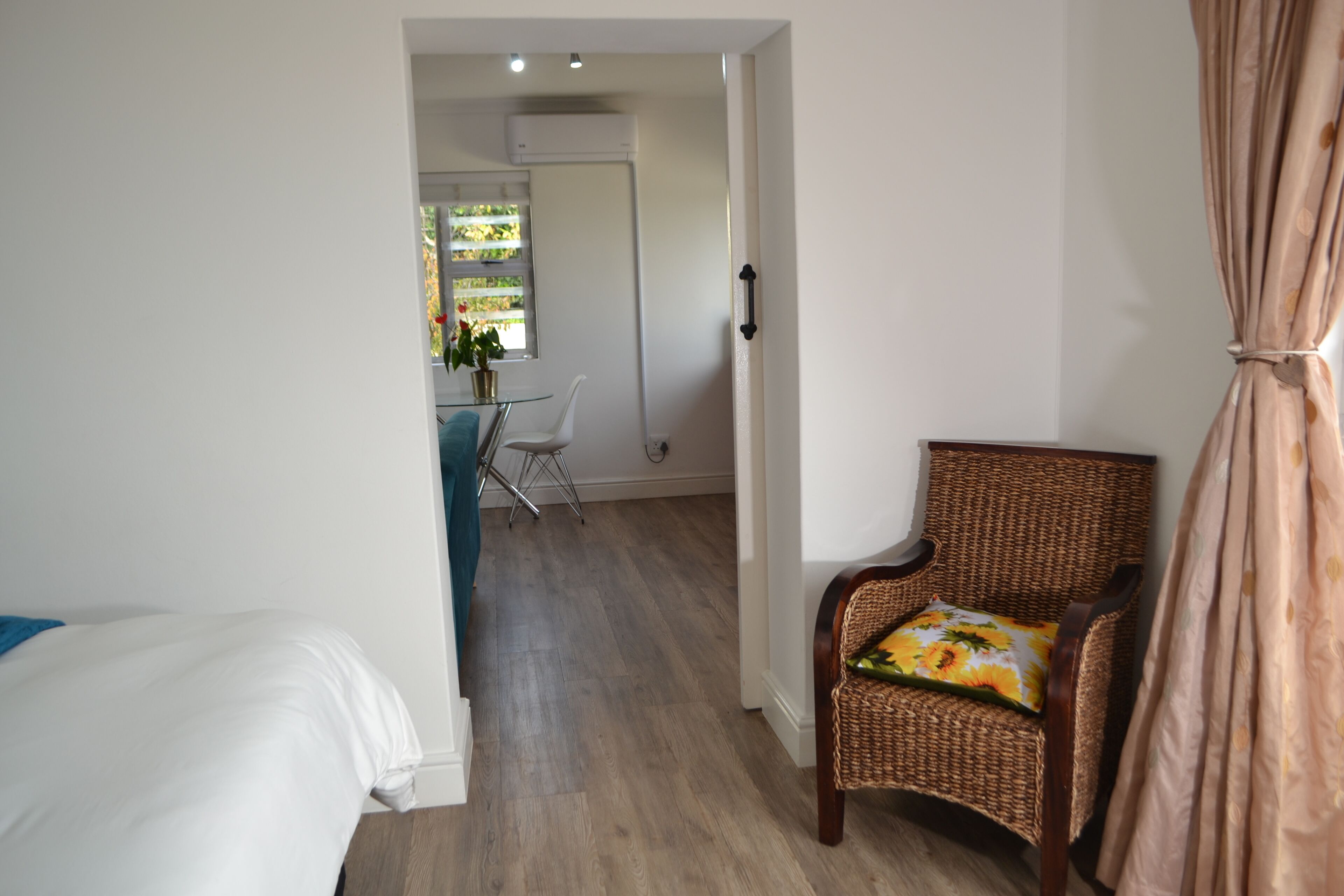 Une pièce comprenant un lit, un fauteuil en osier avec un coussin à motifs floraux, un coin repas avec une table et des chaises, et une fenêtre avec des stores.
