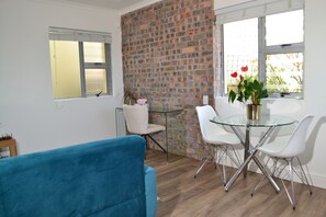 Ein Zimmer mit Ziegelwand, Glastisch mit Stühlen, blaues Sofa und Fenster mit Jalousien.