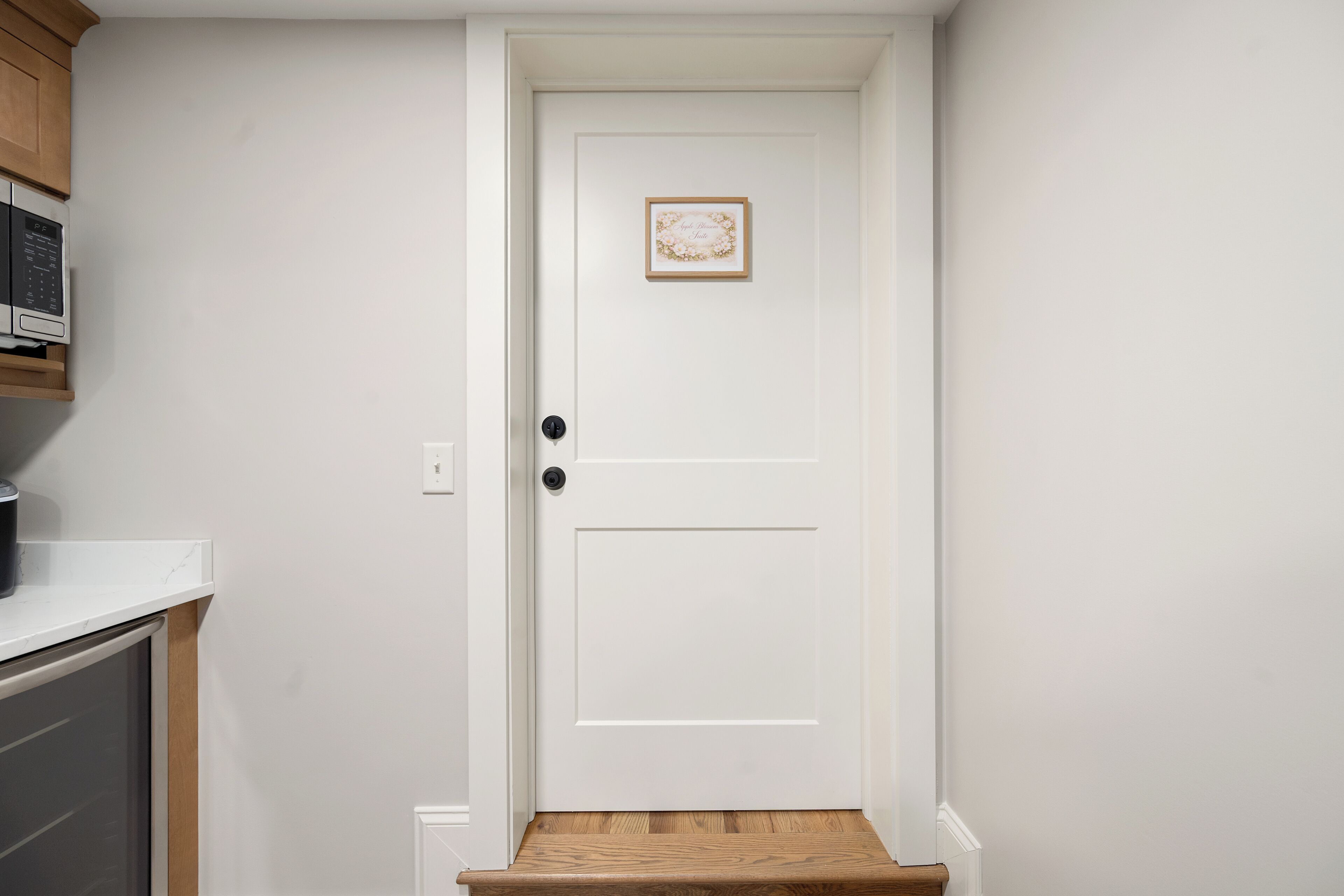 Une porte blanche avec une petite image au-dessus, un four à micro-ondes posé sur un comptoir à gauche, et une marche en bois menant à la porte.