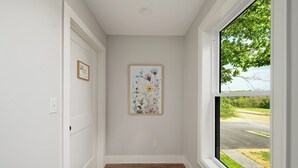 Un pasillo con suelo de madera, un cuadro de flores enmarcado en la pared y una ventana con vistas a una carretera y vegetación.