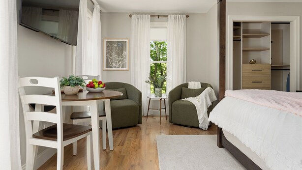 Un dormitorio con una cama, dos sillas, una mesa con fruta y una planta, una ventana con cortinas y un armario.