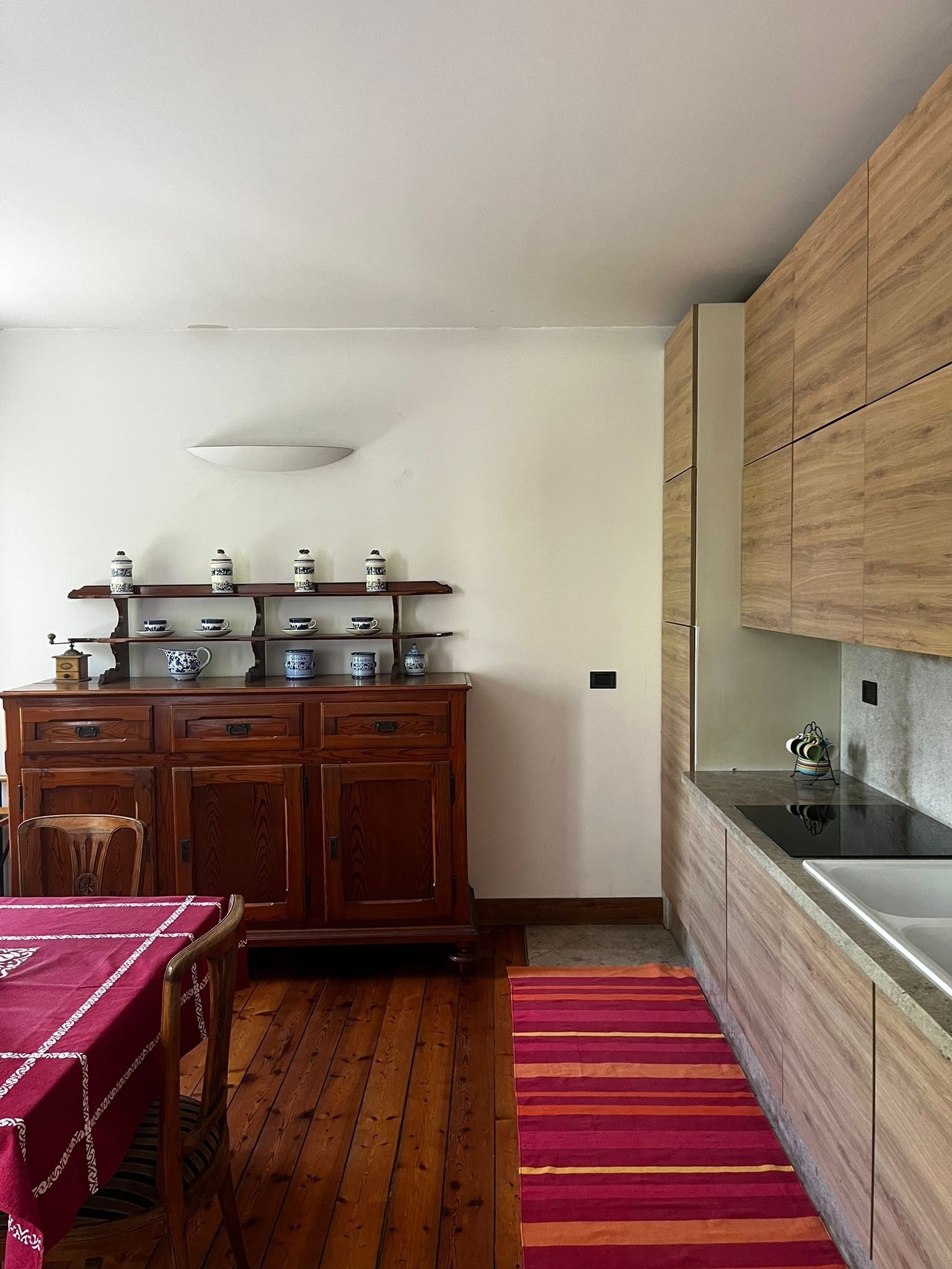 A kitchen with wooden cabinets, a dining table with a red tablecloth, and a striped rug.