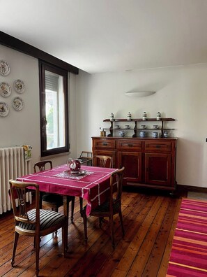 A dining area with a wooden table, chairs, a radiator, a window, and a cabinet with kitchenware. - Historic Palazzo with Pool & Castle View (Sanguinetto)