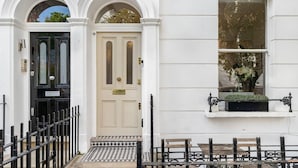 Un bâtiment blanc avec une porte noire, une fenêtre agrémentée d’une jardinière et une clôture en fer noir.