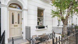Une maison de ville blanche avec une porte à arc traditionnel, un petit espace extérieur aménagé avec une table et des bancs, et une clôture en métal noir qui entoure la propriété.