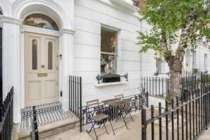 Ein weißes Reihenhaus mit einer traditionellen Bogenförmigen Tür, einer kleinen Außenbereich-Sitzgelegenheit mit Tisch und Bänken sowie einem schwarzen Metallzaun, der das Grundstück umgibt.