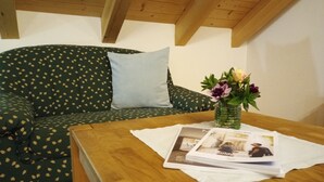 Une pièce avec un plafond en bois, un canapé vert orné de motifs de feuilles, un coussin bleu clair, une table en bois avec un magazine et un vase rempli de fleurs.