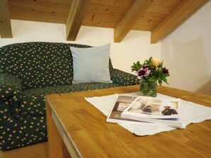 Ein Zimmer mit Holzboden, einem grünen Sofa mit Blättermotiv, einem hellblauen Kissen, einem Holztisch mit einer Zeitschrift und einer Vase mit Blumen.