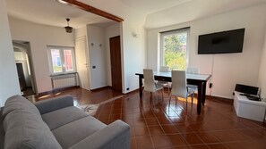 Ein Wohnzimmer mit Sofa, Esstisch mit Stühlen, einem an der Wand befestigten Fernseher und einer durchgehenden Tür erkennbaren Küchenzeile.