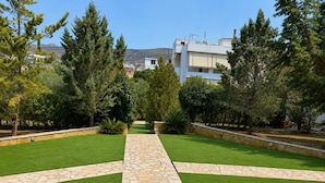 A landscaped garden with a central stone pathway, surrounded by green lawns and trees. - Treasure B B - Garden Stay in Paloukia Salamina (Salamis)