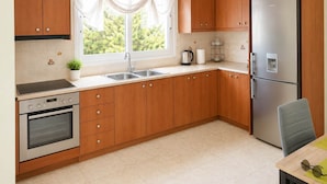 A kitchen with wooden cabinets, a stainless steel refrigerator, a double sink, and a window with a view of greenery. - Treasure B B - Garden Stay in Paloukia Salamina (Salamis)