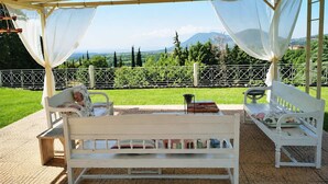 A covered outdoor seating area with white benches and a table, overlooking a green landscape with trees and mountains. - Studio Aspilon Pool and Mountain Views Near Drama (Drama)