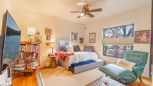 Un dormitorio con cama, una silla, estantería, ventilador de techo y ventana con persianas.