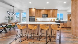 A modern kitchen with wooden cabinets, a central island, bar stools, and a dining area with a table and chairs. - Reno Retreat - 3BR Oasis w Swim Spa Hot Tub (Reno)