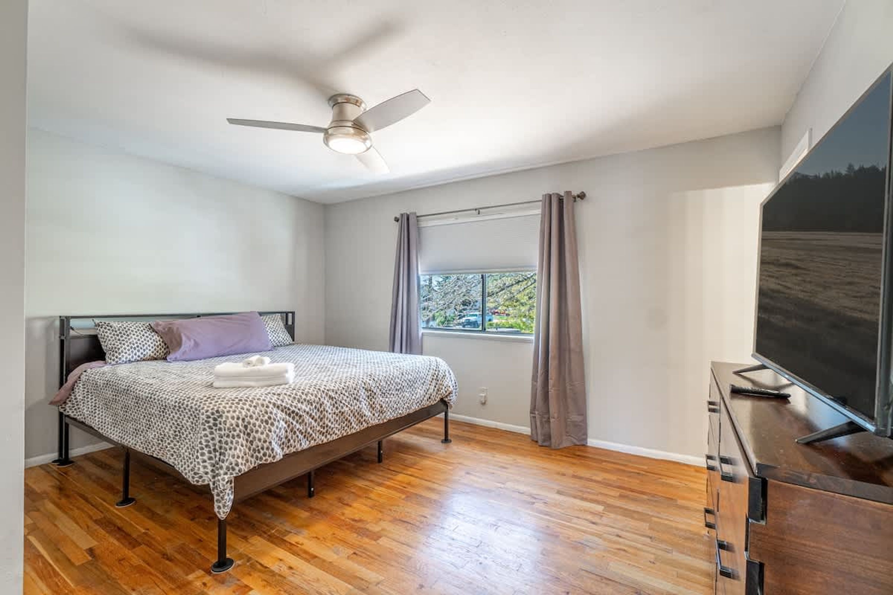 A bedroom with a bed, a ceiling fan, a window with curtains, and a TV on a wooden cabinet.