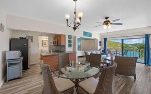 A modern kitchen with a refrigerator, microwave, and a cart, a dining area with wicker chairs and a glass table, and a balcony with a view of the landscape.