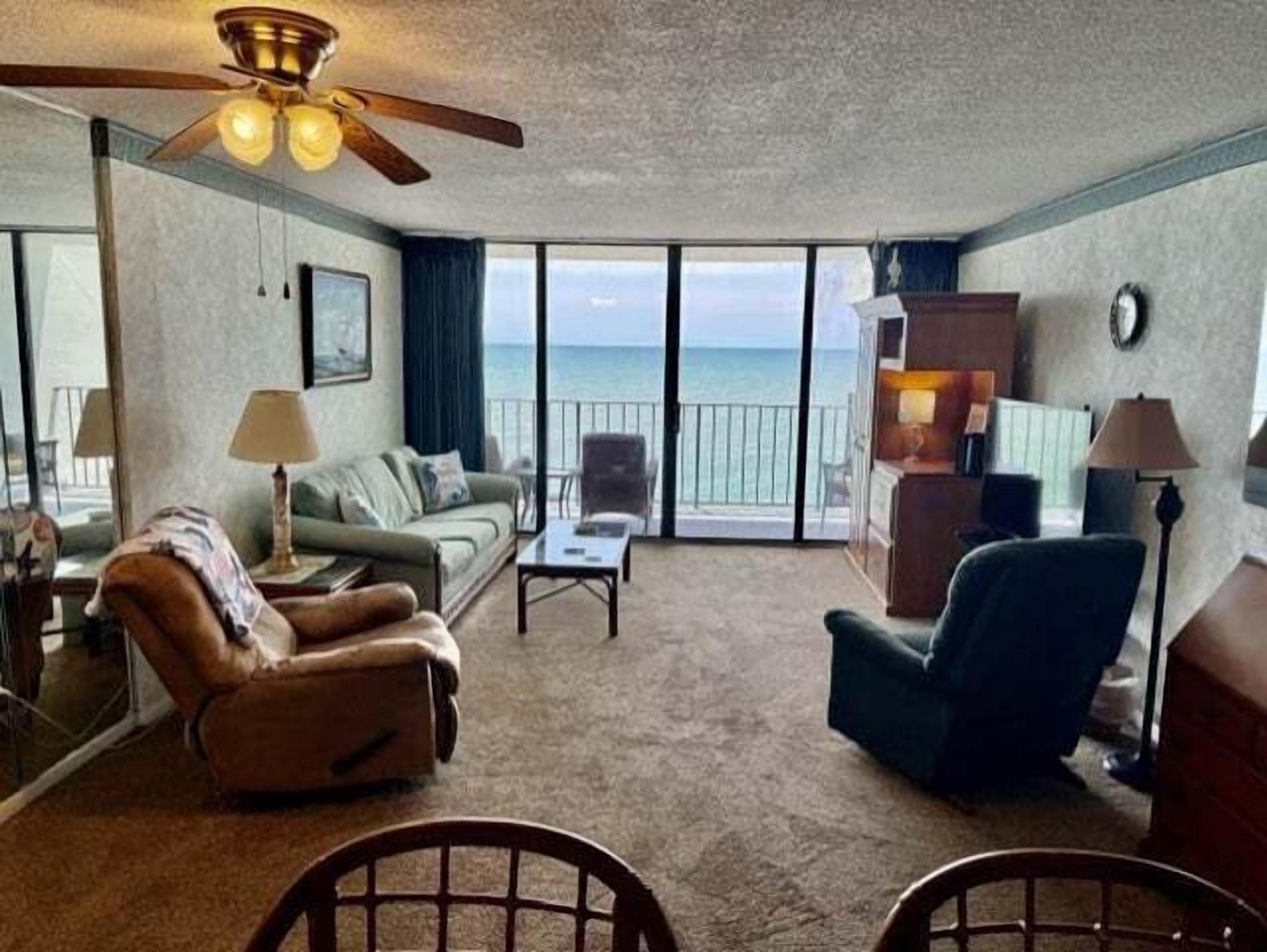 A hotel room with a balcony, a ceiling fan, a sofa, a chair, and a small table.