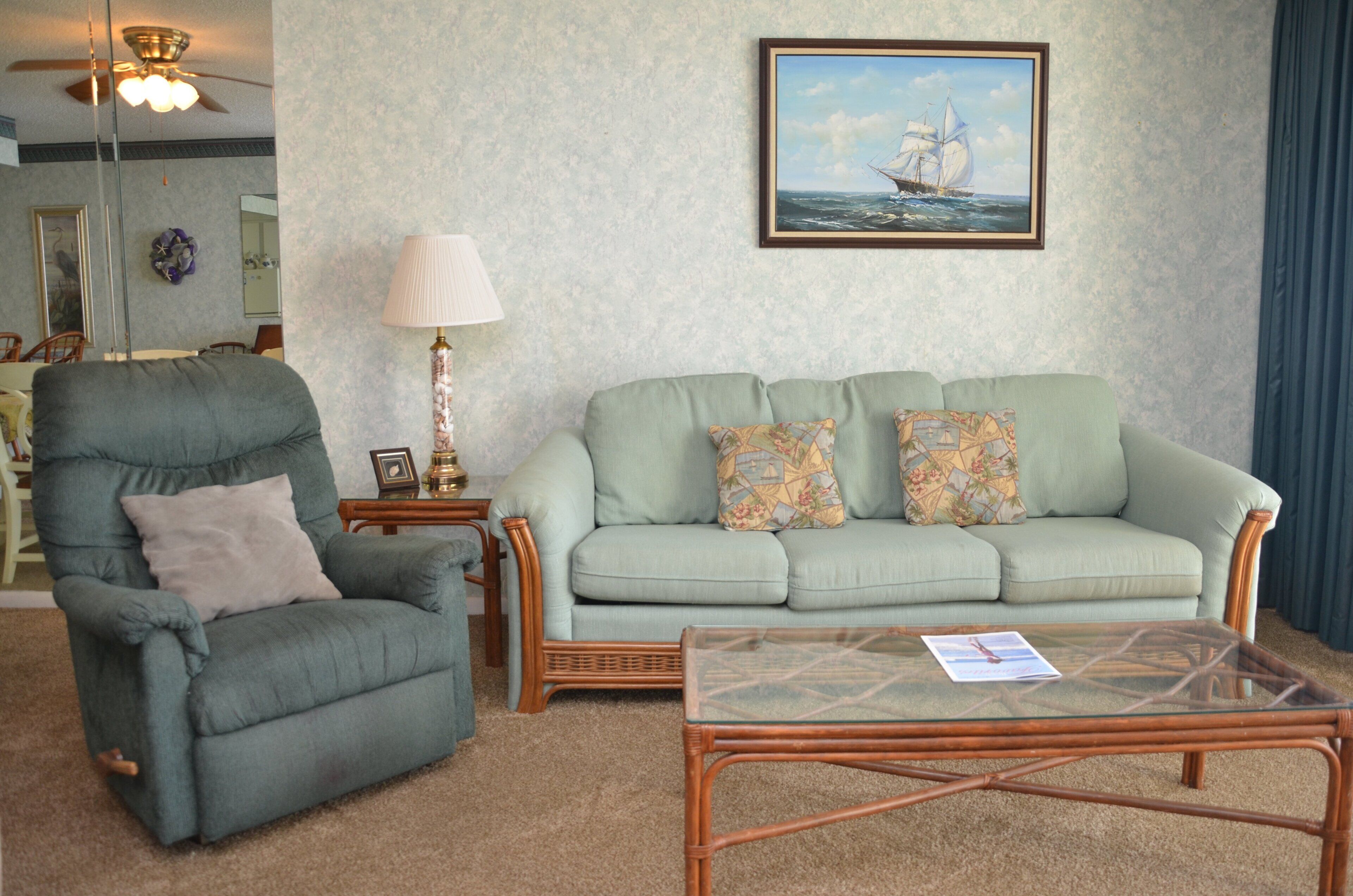 A living room with a green armchair, a light green sofa, a glass coffee table, a lamp, and a framed picture on the wall.
