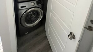A compact laundry room with a front-loading washer and dryer, a door with a silver handle, and a wooden floor. - Comfortable Private Room – Central Winnipeg (Winnipeg)