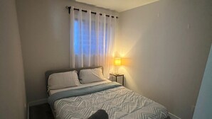 A single bed with a patterned bedspread, a bedside lamp, and sheer curtains. - Comfortable Private Room – Central Winnipeg (Winnipeg)