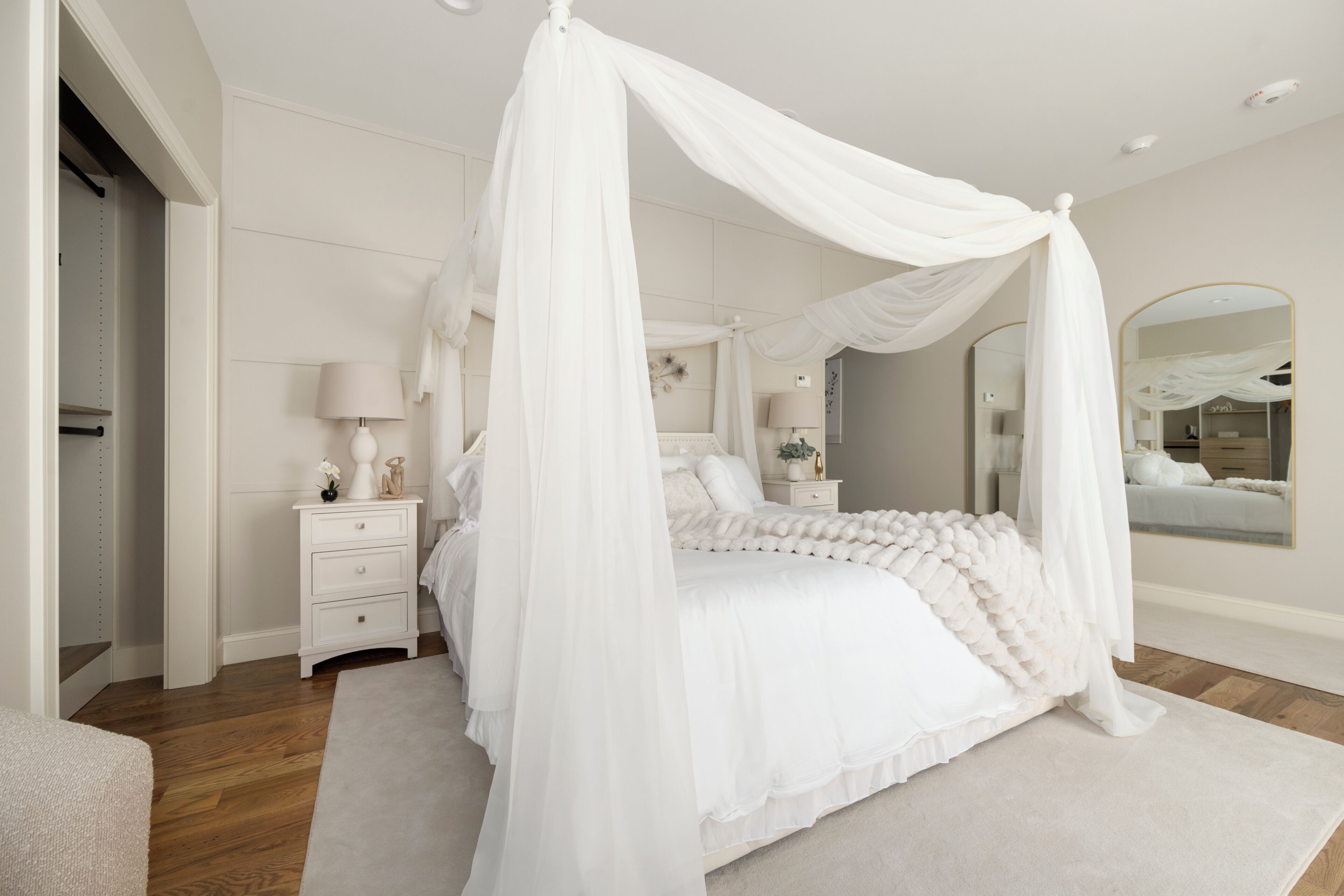 A bedroom with a four-poster canopy bed, a bedside table with a lamp, a mirror, and a wardrobe.