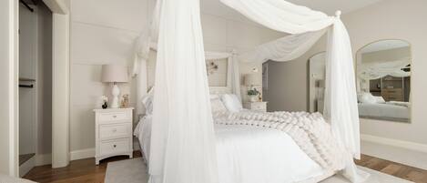 A bedroom with a four-poster canopy bed, a bedside table with a lamp, a mirror, and a wardrobe.