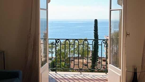Una porta oberta que dona a un balcó amb vista al mar i un arbre allunyat.