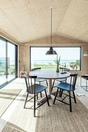 Un espace repas comprenant une table ronde, des chaises, un luminaire suspendu et une vue sur la mer grâce à de grandes fenêtres.